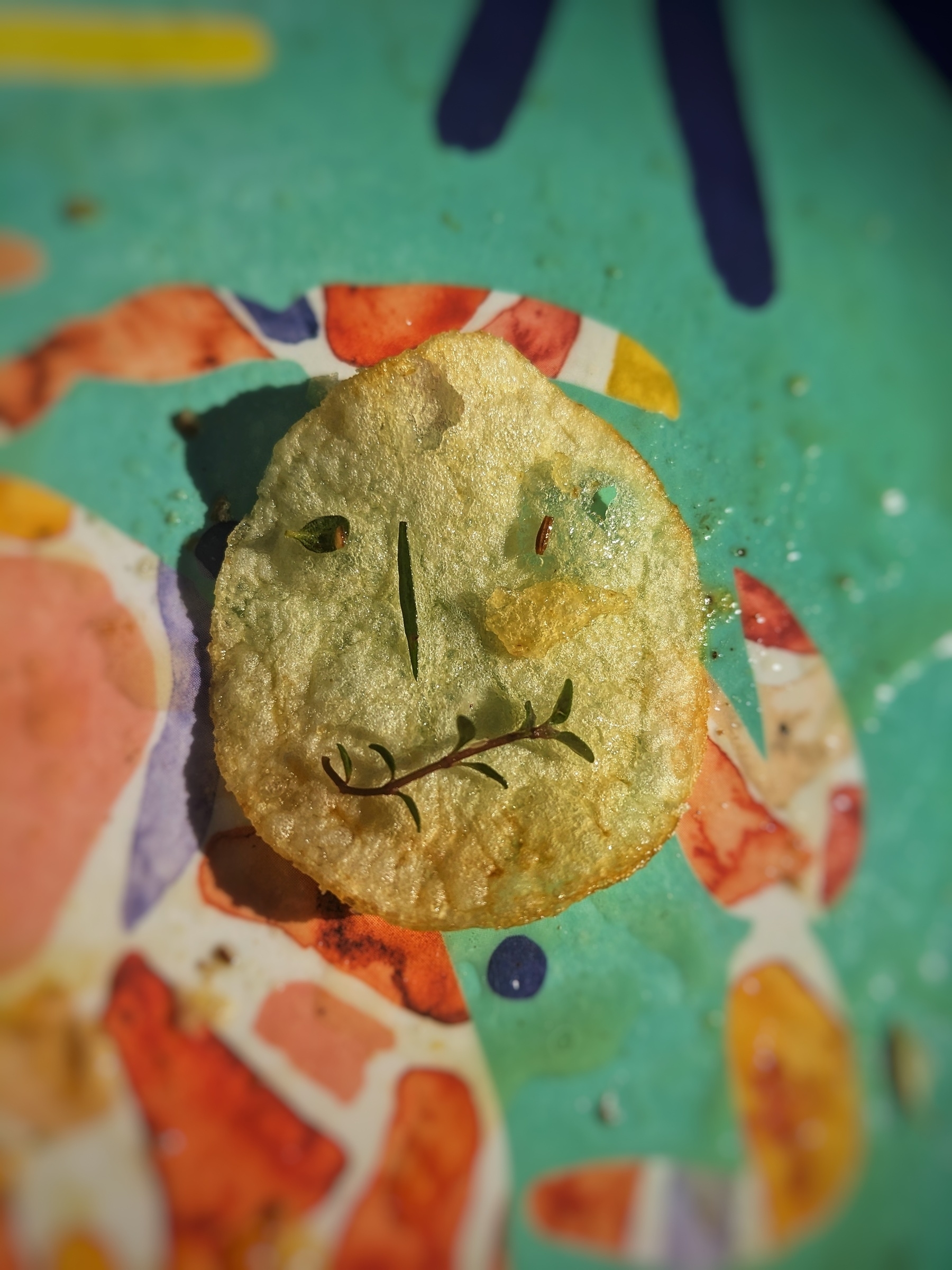 Auto-generated description: A potato chip is creatively arranged with herbs to resemble a face on a colorful background.