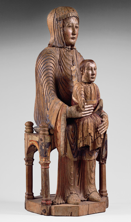 A wooden sculpture depicts a seated Madonna holding a smaller Christ on her lap, both adorned in draped garments.