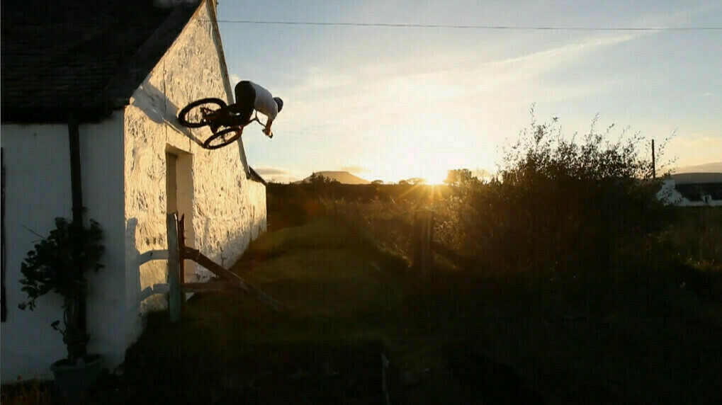 Semi-auto-generated description: Danny Macaskill is performing a wall ride on a bicycle against a white building at sunset.