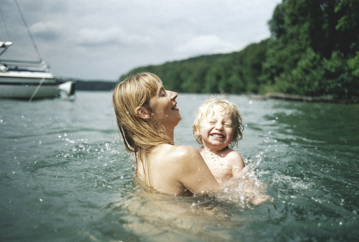 Auto-generated description: A woman and a child are joyfully swimming in a lake near a boat and a forested area.