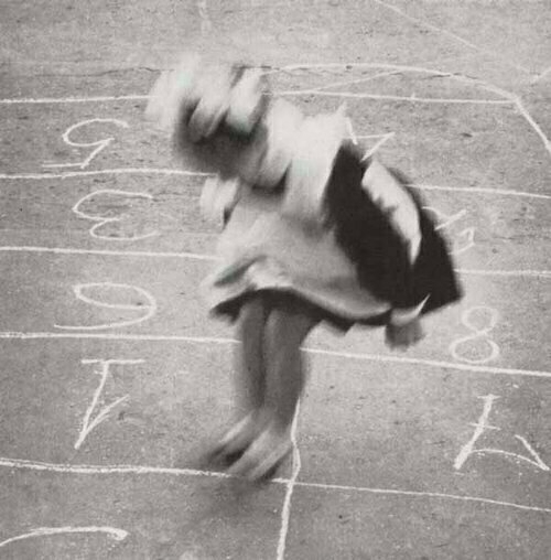 Auto-generated description: A girl from the late 19th or early 20th C is playing hopscotch on a chalk-drawn grid.