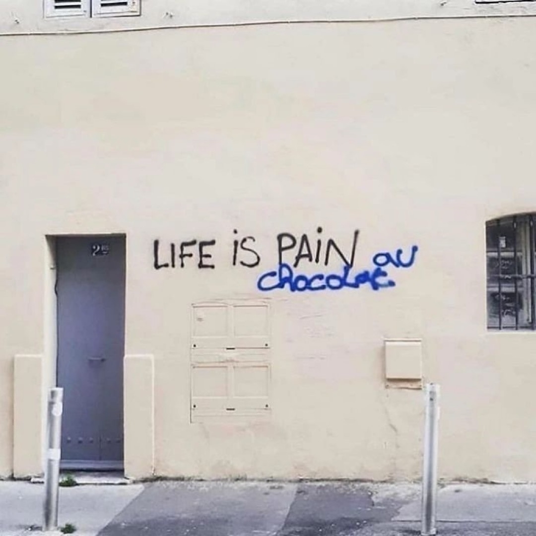 Auto-generated description: Graffiti on a wall reads LIFE IS PAIN au chocolat with the words au chocolat humorously added to the original message.