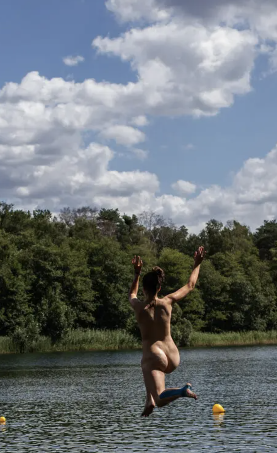 Auto-generated description: A person is joyfully jumping into a lake surrounded by trees under a partly cloudy sky.