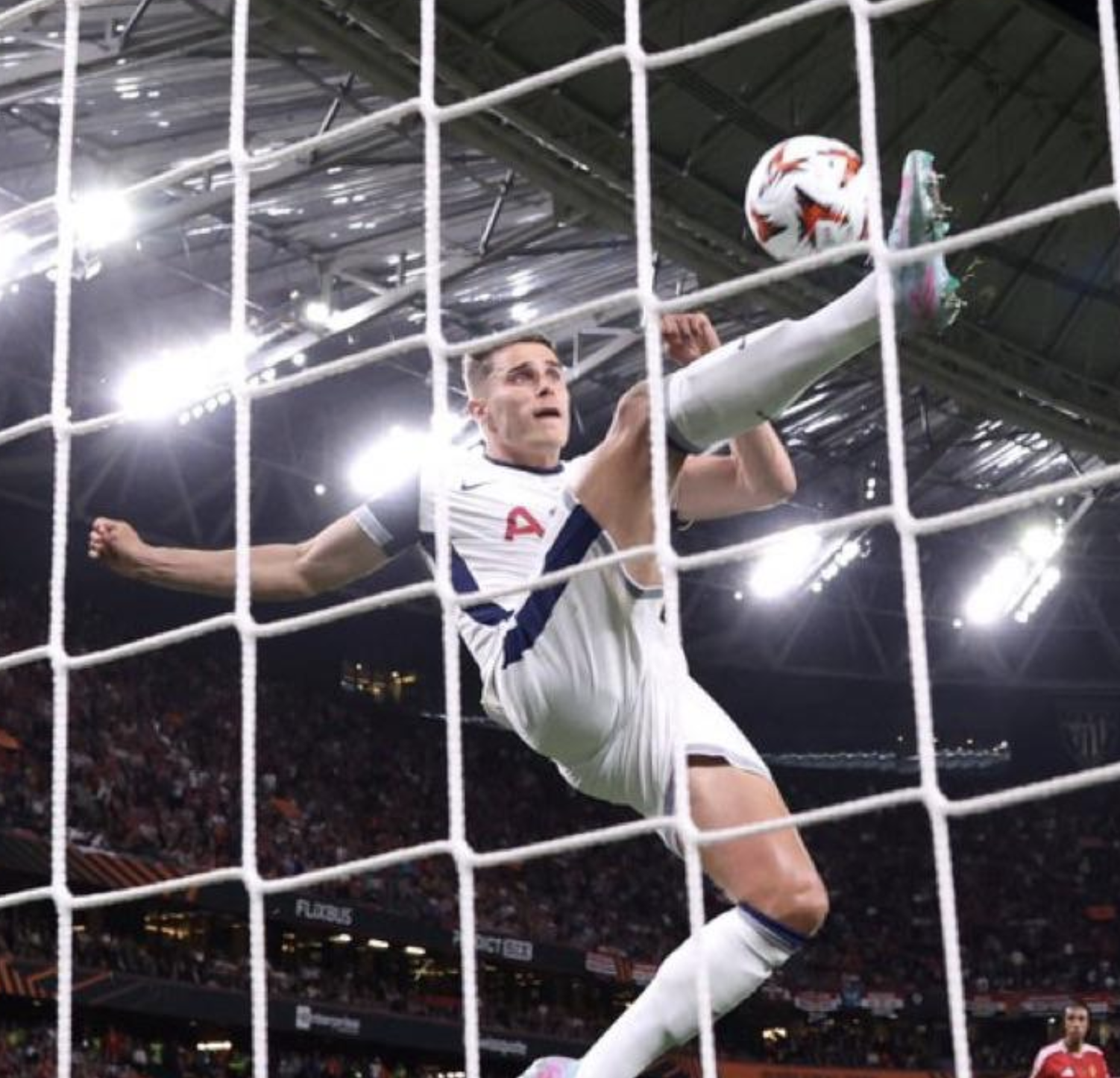 Auto-generated description: A soccer player in a white and navy kit performs an acrobatic kick near the goal net in a stadium.
